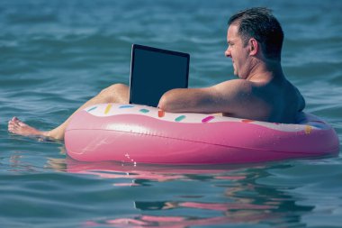 Her yerde ve her zaman çalışmak. Sudaki şişme halka üzerinde dizüstü bilgisayar kullanan genç bir adam çalışıyor. Serbest çalışma, tatiller, mesai, sosyal mesafe