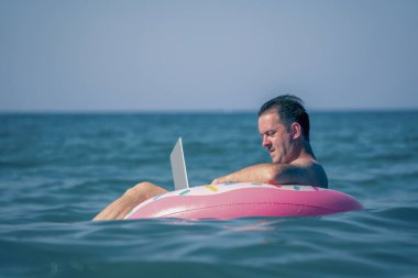 Her yerde ve her zaman çalışıyorum. Sudaki şişme halka üzerinde dizüstü bilgisayar kullanan bir adamın portresi.. 