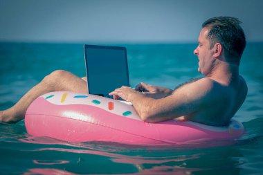 İşadamı çalışıyor. Sudaki şişme halkada dizüstü bilgisayar kullanıyor. Serbest çalışma, tatiller, mesafe çalışması, sosyal uzaklık, e-öğrenme, bağlantı kavramı. 