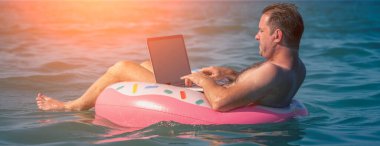 Sudaki şişme halka üzerinde dizüstü bilgisayar kullanan genç bir adam çalışıyor. Serbest çalışma, tatiller, mesafe çalışması, sosyal uzaklık, e-öğrenme, bağlantı, yaratıcı profesyonellik, online konsept buluşması