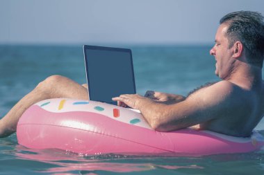 Sudaki şişme halka üzerinde dizüstü bilgisayar kullanan genç bir adam çalışıyor. Serbest çalışma, tatiller, mesafe çalışması, sosyal uzaklık, e-öğrenme, bağlantı kavramı. 