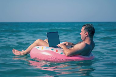 Kavramsal görüntü: Her yerde ve her zaman çalışabilirsiniz! Diz üstü bilgisayarı olan bir adam deniz ya da okyanus suyunda şişirilebilir bir halka üzerinde