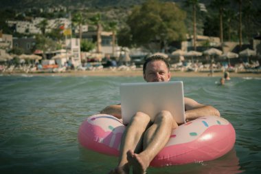 Her yerde ve her zaman çalışmak. Deniz suyunda şişme halkası olan serbest çalışan bilgisayar üzerinde çalışır. Suda bacakları olan ve gümüş dizüstü bilgisayar kullanan mutlu bir adam..