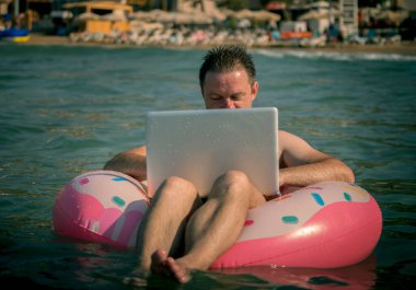 Deniz suyunda şişme halkası olan serbest çalışan bilgisayar üzerinde çalışır. Suda bacakları olan ve gümüş dizüstü bilgisayar kullanan mutlu bir adam.. 