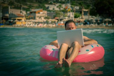 Deniz suyunda şişme halkası olan serbest çalışan bilgisayar üzerinde çalışır. Gümüş dizüstü bilgisayar kullanarak yüzme çemberindeki mutlu adam. Boşluğu kopyala.