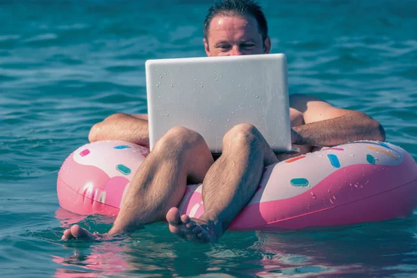 Genç adam dizüstü bilgisayarda deniz suyunda şişme halkayla çalışıyor. Boşluğu kopyala