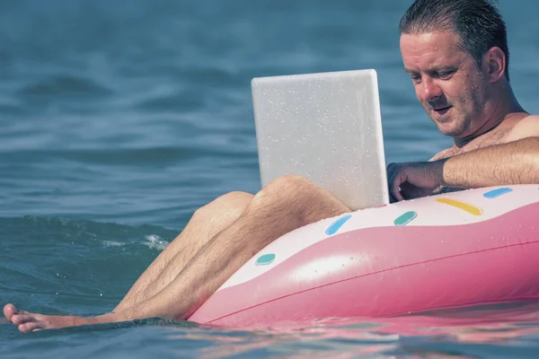 Tatilde çalışan erkek girişimciyi deniz ve okyanus sularında şişirilebilir halka üzerine dizüstü bilgisayarla kapatın. Yatay resim.
