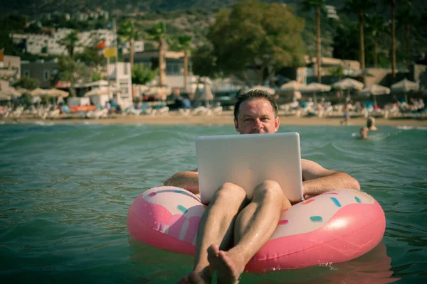 Her yerde ve her zaman çalışmak. Deniz suyunda şişme halkası olan serbest çalışan bilgisayar üzerinde çalışır. Suda bacakları olan ve gümüş dizüstü bilgisayar kullanan mutlu bir adam..