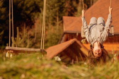 Genç ve güzel bir kız baş aşağı asılı ve dışarıda salıncakta eğleniyor. Mutlu çocukluk, yaz tatili, tatil ve dinlenme konsepti. Tasarım için alanı kopyala.