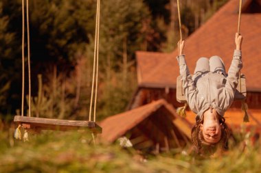 Genç ve güzel bir kız baş aşağı asılı ve dışarıda salıncakta eğleniyor. Mutlu çocukluk, yaz tatili, tatil ve dinlenme konsepti. Yatay görüntü. Tasarım için alanı kopyala.