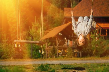 Genç ve güzel bir kız baş aşağı asılı ve dışarıda salıncakta eğleniyor. Mutlu çocukluk, yaz tatili, tatil ve dinlenme konsepti. Yatay resim.
