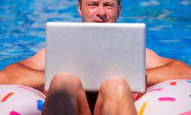 Bu yaz çalışanları ofisten nasıl çıkaracağız? İş adamı yüzme havuzunda şişme yüzükle bilgisayarla uzaktan çalışıyor. Yatay resim.