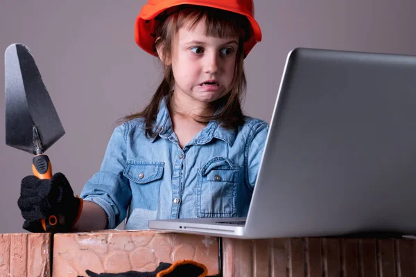Humorous image of confused and scared cute little builder with laptop builds a brick wall. Building and repair services concept.