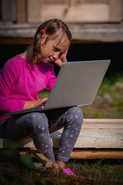 Conceptual photography: Learn always and everywhere. Portrait of young beautiful school girl uses a laptop and studies remotely outdoorson. Vertical image.