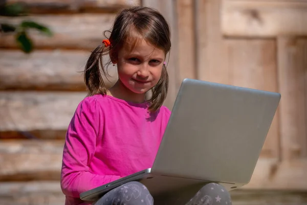 Conceptual photography: Learn always and everywhere. Portrait of young beautiful school girl uses a laptop and studies remotely outdoorson the background of a wooden house.