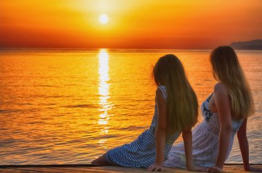 Anne ve kızının deniz kıyısındaki bir köprüde oturup sabah güneşiyle buluşmalarını. Yaz tatili, dinlenme, iletişim ve seyahat konsepti.