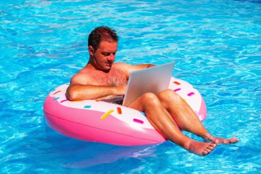 Yüzme havuzunda şişme bir dairede oturan ve bilgisayarla uzaktan çalışan iş adamının portresi. Yatay resim.