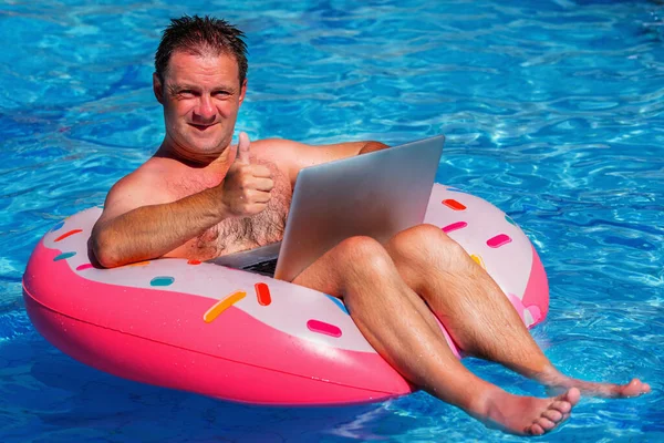 Portrait of businessman during summer vacation on an inflatable circle in the pool works remotely with laptop. Horizontal image.