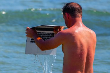 Dizüstü bilgisayarına iyi bakmanın zamanı geldi. Deniz kenarında yaz tatili yapan bir adamın komik görüntüsü dizüstü bilgisayarını suda yıkar..