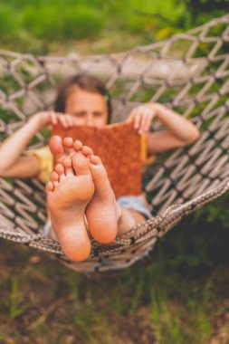 A young beautiful girl lies in a hammock and reads a book. Rest, summer vacation, leisure time concept. Selective focus on feets.