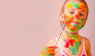 Küçük tatlı kız fırça ve renkli boyalı ellerle kendine çocuk makyajı yapıyor. Güzellik, eğlence, mutlu çocukluk ve sanat konsepti. Tasarım veya metin için boşluk kopyala.