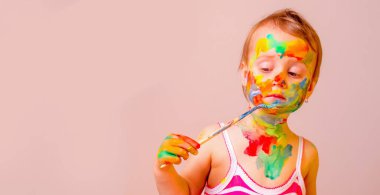 Küçük tatlı kız fırça ve renkli boyalı ellerle kendine çocuk makyajı yapıyor. Güzellik, eğlence, mutlu çocukluk ve sanat konsepti. Boşluğu kopyala.