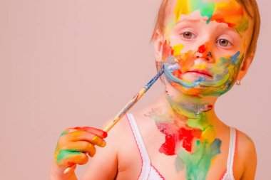 Küçük tatlı kız fırça ve renkli boyalı ellerle kendine çocuk makyajı yapıyor. Güzellik, eğlence, mutlu çocukluk ve sanat konsepti. Yatay resim.