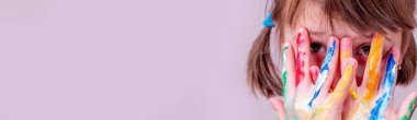 Portrait of really happy little cute child girl looking throught fingers. Selective focus on eyes. Colorful painted hands as symbol of joyful happy life. Copy space.