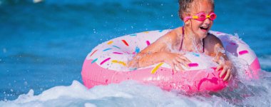 Joyful child girl on inflatable ring riding on breaking wave. Travel, healthy lifestyle and summer holiday concept. Copy space for text.