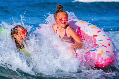Mutlu çocuklar sahilde sörf yapıyor. Şişme yüzüklerin üzerindeki neşeli çocuklar dalgaları kırıyor. Seyahat, sağlıklı yaşam tarzı, yüzme aktiviteleri ve yaz tatili konsepti..