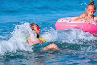 Mutlu çocuklar sahilde sörf yapıyor. Şişme yüzüklerin üzerindeki neşeli çocuklar dalgaları kırıyor. Seyahat, sağlıklı yaşam tarzı, yüzme aktiviteleri ve yaz tatili konsepti. Tropikal adada tatiller
