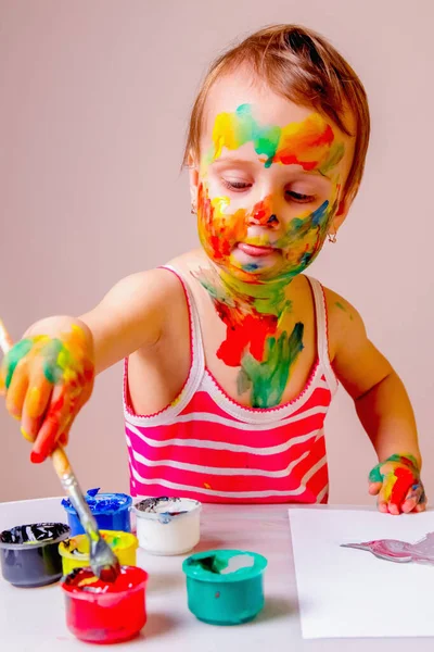 Painting lessons. Portrait of little cute girl with children\'s colorful makeup paints the picture. Art, education and childhood concept. Vertical image.