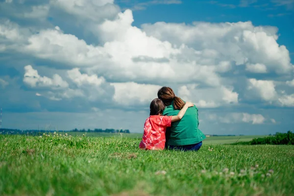 Back view of two young girls sisters hug each other in the green field against beautiful background of wild nature and cloudy sky. Love, happy childhood concept. Horizontal image.
