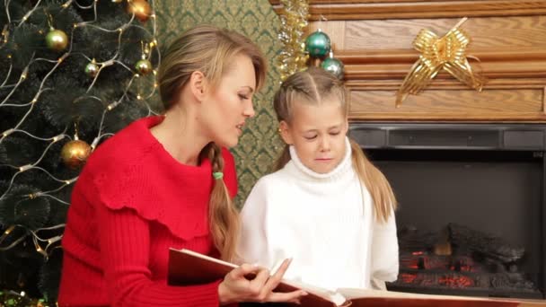 Madre leyéndole un cuento a su hija bajo un árbol de Navidad. Leer una historia . — Vídeo de stock