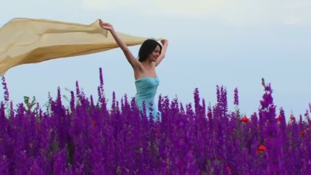 Meisje met de ontwikkeling van de sjaal. mooi meisje loopt door een veld van mooie bloemen groeien in de wind met haar sjaal. bewegende camera. — Stockvideo