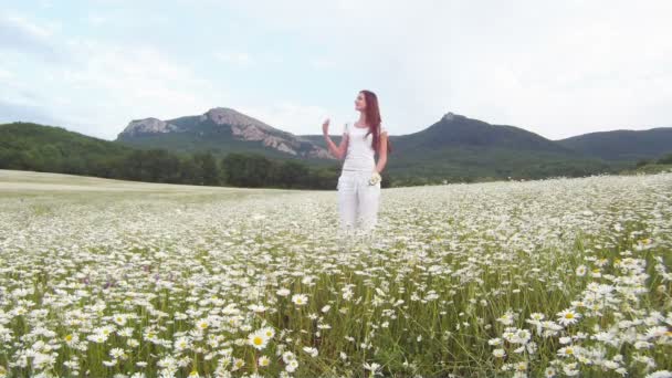 她走穿过田野。美丽的女孩在白色的礼服走上洋甘菊字段的山区地形背景. — 图库视频影像