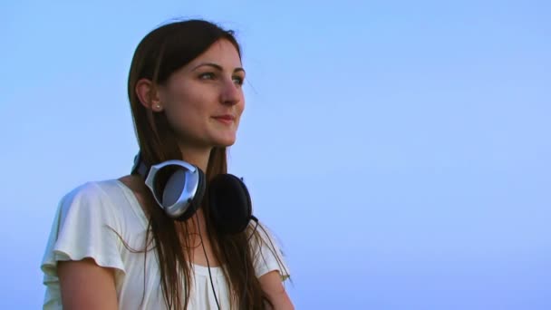 Dziewczyna w słuchawkach na panoramę. nosi duże słuchawki i słuchania muzyki w tle piękne błękitne niebo. — Wideo stockowe