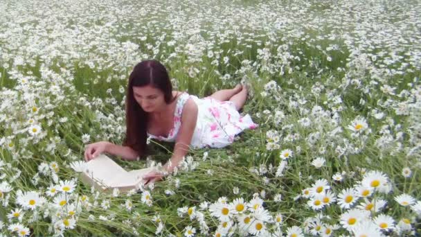 Rapariga a ler um livro. Uma jovem se senta em um belo campo de camomila e lê mais livros . — Vídeo de Stock