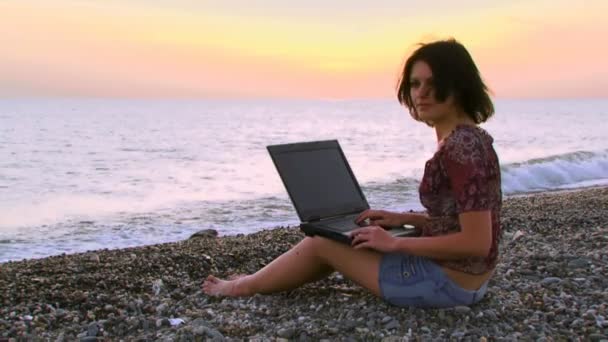 Chica con un portátil en el mar. Una hermosa chica sentada en la playa y trabajando en el portátil . — Vídeos de Stock