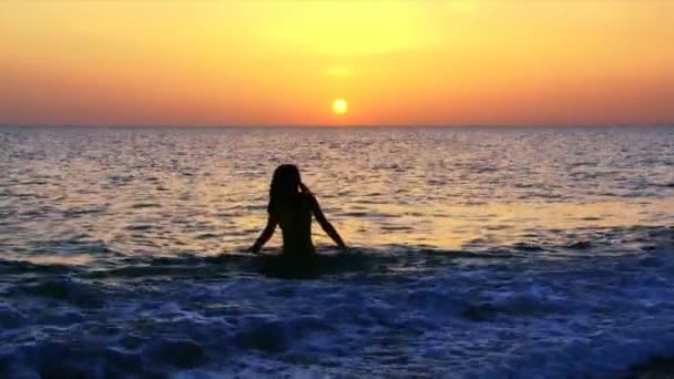 Sunset at sea. A girl stands on the beach watching the sunset. The waves wash her feet. — Stock Video