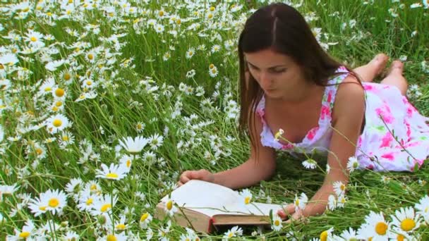 Jovencita leyendo un libro. Una joven se sienta en un hermoso campo de manzanilla y lee más libros . — Vídeos de Stock