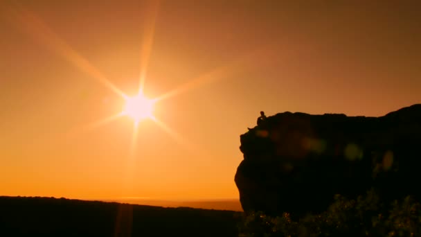 美しい太陽。シルエット。男は崖の端に座っています。熱狂的に彼の手を発生させます. — ストック動画