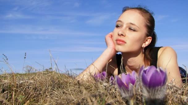 A rapariga está na natureza. Uma mulher bonita encontra-se na natureza perto de flores roxas. Contra o fundo do céu azul . — Vídeo de Stock