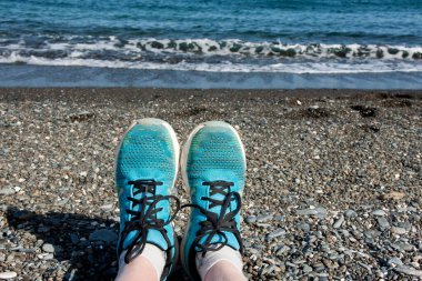 Denizin arka planına karşı çakıl taşı plajında mavi spor ayakkabılı kadınların bacakları. Deniz kıyısında spor ve rahatlama. Akdeniz, Türkiye, Antalya, Kemer.