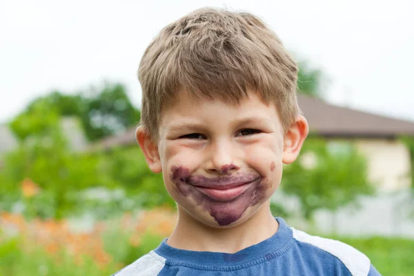 Close-up portrait of child whose face is stained with black mulberry juice. Smiling preschooler boy stained his face in lilac. Happy childhood. Summer. Time to eat berries.