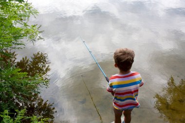 preschool child is fishing in river on cloudy summer day. boy threw fishing rod, stands with his back to camera, clouds are reflected in river. Nature. Outdoor. Happy and interesting childhood