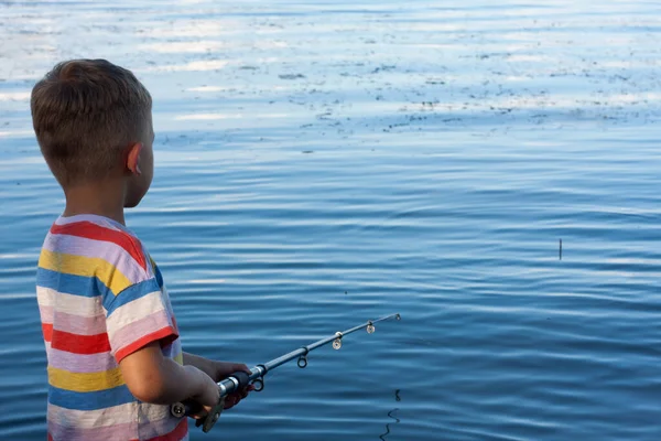 preschool child is fishing in river on summer day. boy has thrown bait and is waiting for bite, standing with his back to camera. Amateur fishing in open water. Nature. Happy childhood. copy space