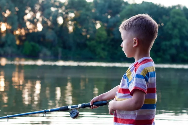 Portrait of boy with fishing rod in profile. Focused young fisherman is fishing in river on summer day. Amateur fishing in open water from river bank. Sunset. Nature. Happy and interesting childhood