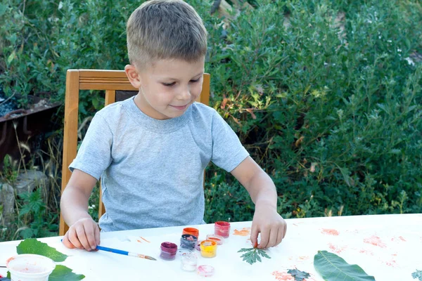 child paints leaves with paints, draws a picture, making prints of leaves. Childrens creativity in nature. Distance learning. Outdoor. Summer.