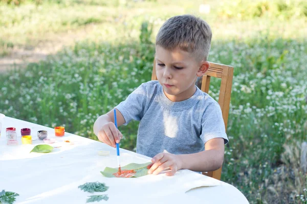 child paints leaves with paints, draws a picture, making prints of leaves. Childrens creativity in nature. Distance learning. Outdoor. Summer.